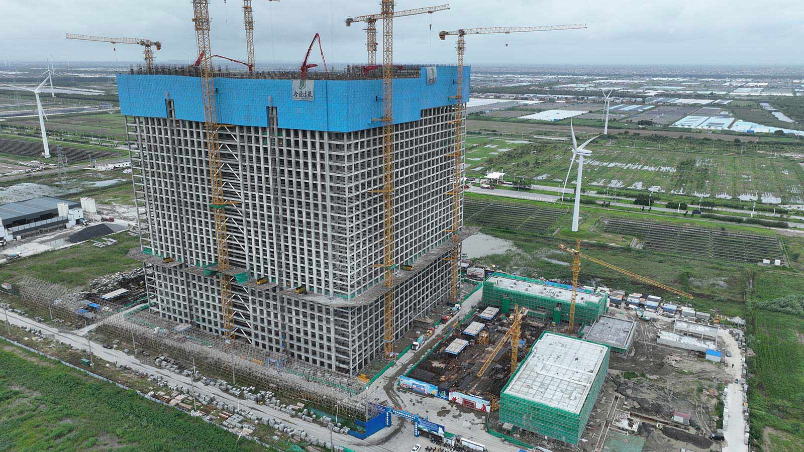 Very large cube-shaped building, about 100 metres per side, under construction, with wind turbines on flat land in the background
